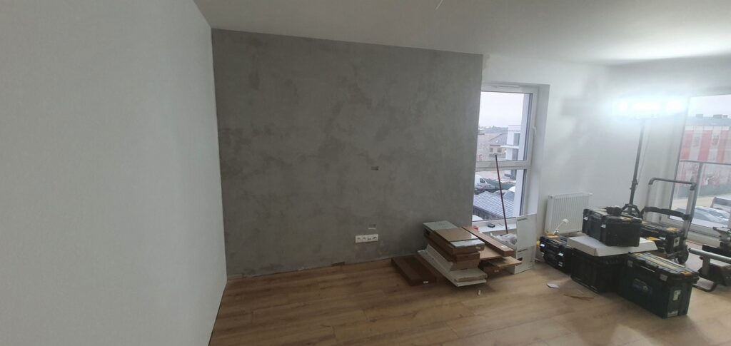 Przywidz - Remont wnętrz w mieszkaniu, z ukończoną ścianą pokrytą tynkiem oraz świeżo położoną podłogą. W tle widoczne materiały i narzędzia używane przez firmę remontową.
