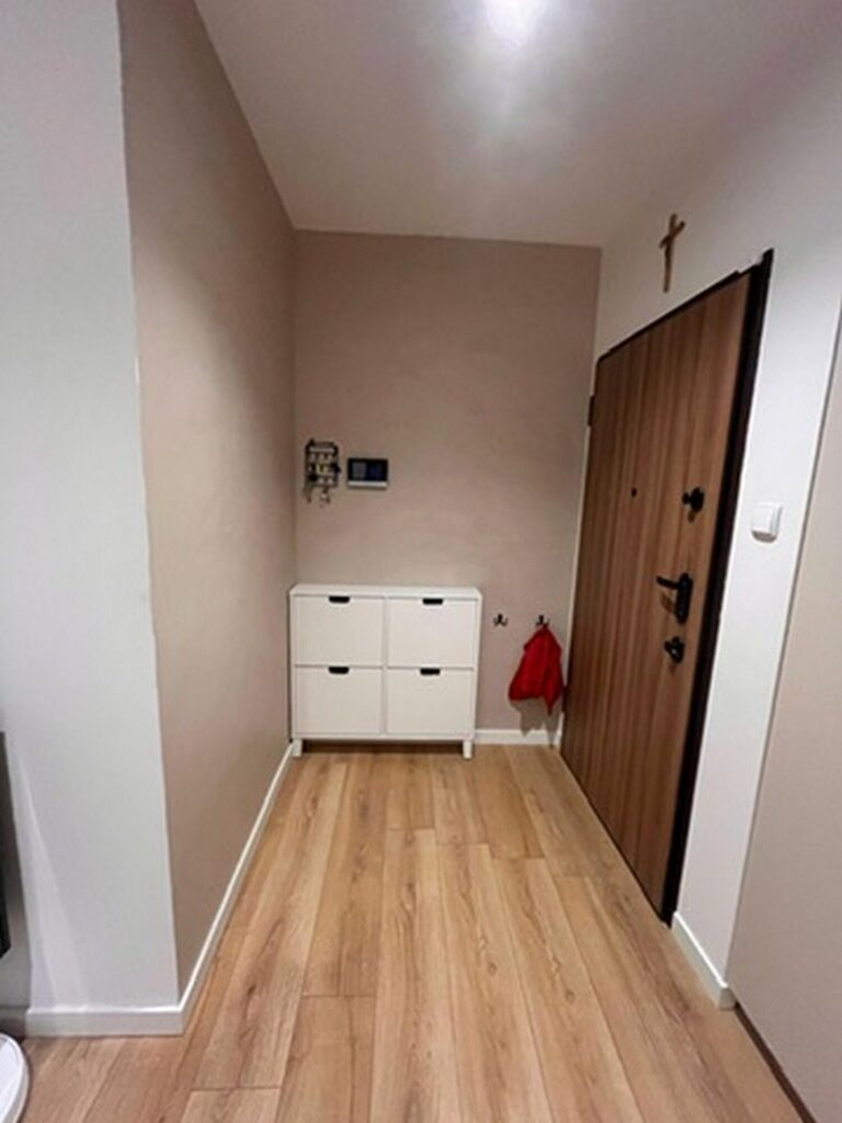 Nowoczesny korytarz po remoncie, z eleganckimi wykończeniami wnętrz, stworzony przez firmę remontową. Widoczna jasna ściana i drewniana podłoga, które nadają przestrzeni przytulny charakter.