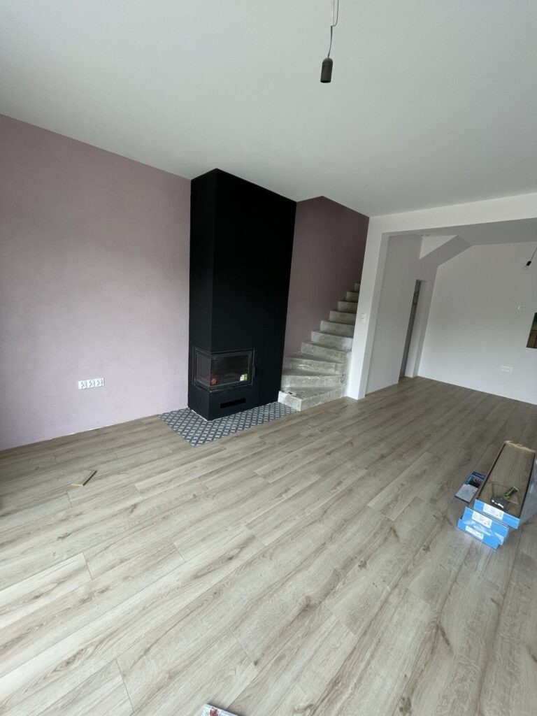Nowoczesny salon po remoncie z przytulnym kominkiem i minimalistycznymi wykończeniami wnętrz. Widać świeże prace firmy remontowej oraz elegancką podłogę.