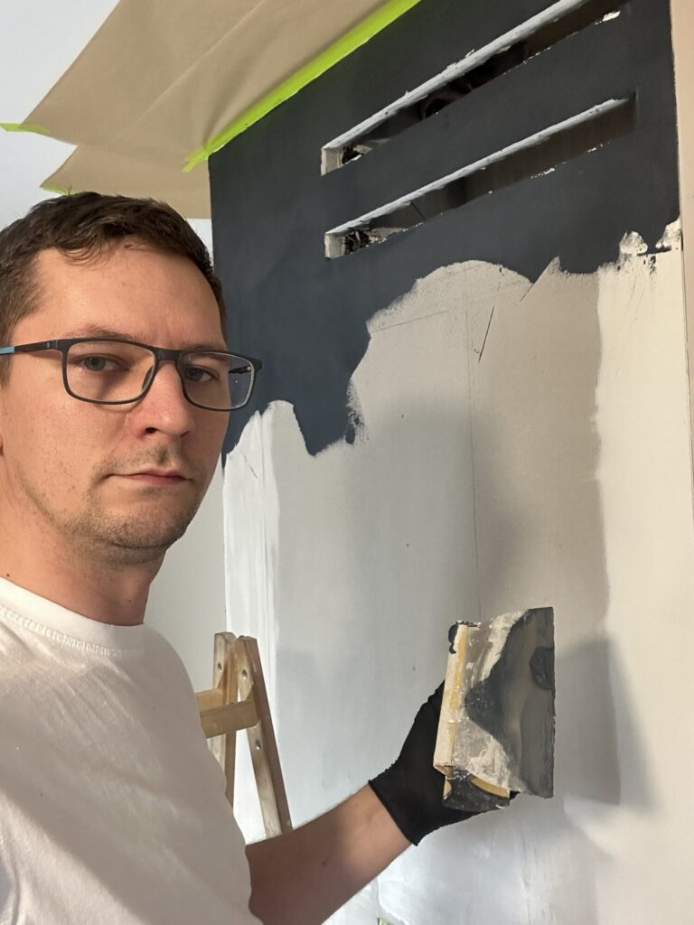 Mężczyzna przy pracy nad remontem wnętrza, malując ścianę na czarno. W tle widoczna drabina i przygotowania do wykończenia wnętrz. Pracownik firmy remontowej używa narzędzi malarskich.