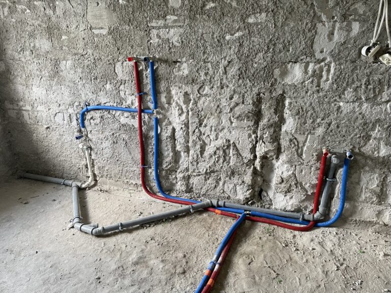 Instalacja hydrauliczna w trakcie remontu mieszkania, przygotowywana przez firmę remontową. Widać różnokolorowe rury z wodą, co sugeruje prace wykończeniowe wnętrz.