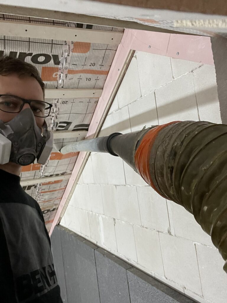 Mężczyzna w maseczce pracujący nad remontem wnętrza, wykorzystujący sprzęt do wykończeń. W tle widoczne ściany przygotowane do malowania, co wskazuje na usługi firmy remontowej.