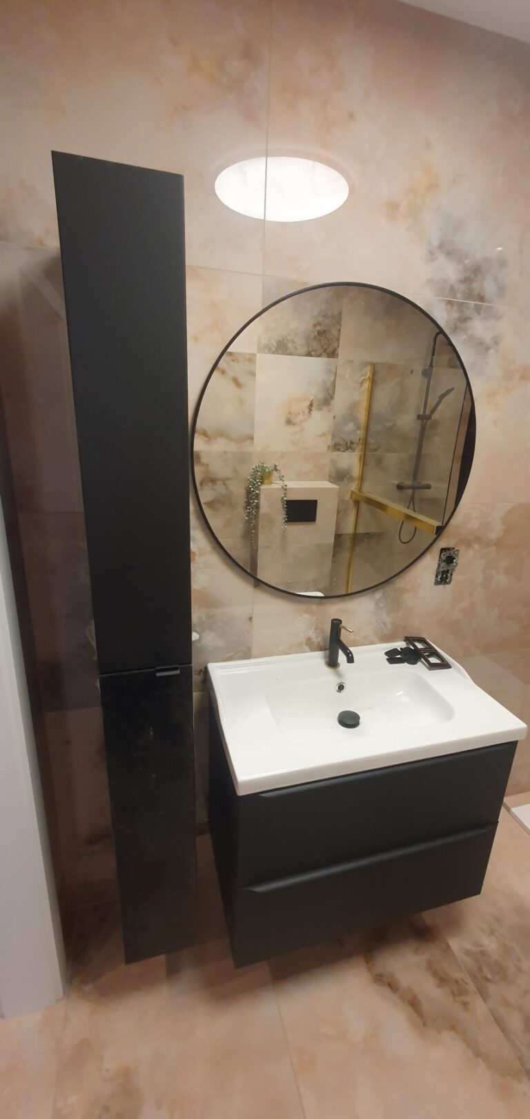 Nowoczesna łazienka po remoncie z eleganckimi wykończeniami wnętrz, zawierająca czarną szafkę, okrągłe lustro i złote akcenty. Idealne rozwiązanie od firmy remontowej.