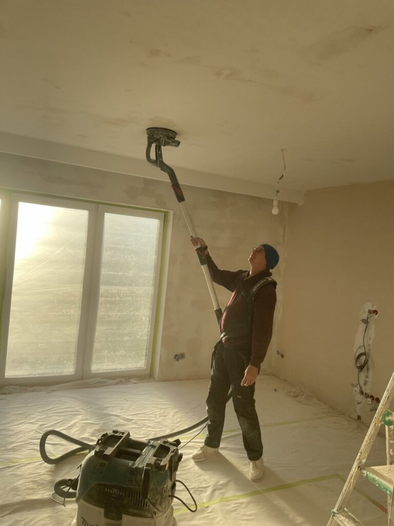 Pracownik firmy remontowej wykonuje wykończenia wnętrz, używając maszyny do szlifowania sufitów w świeżo remontowanym pomieszczeniu.