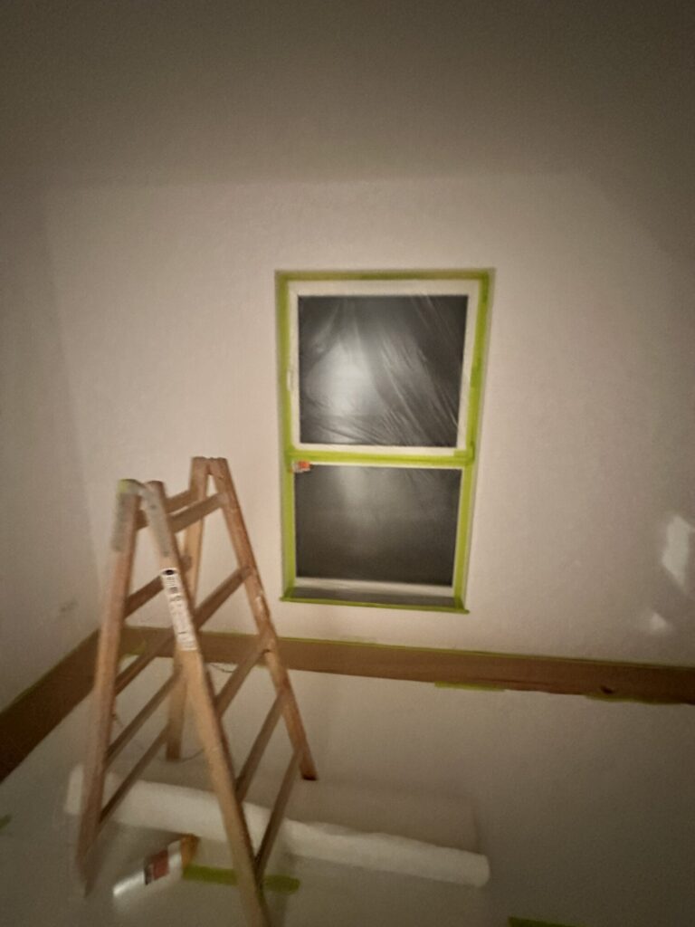 Remont wnętrza z drabiną i oknem zabezpieczonym folią. Prace wykończeniowe w toku, widoczne akcesoria remontowe.