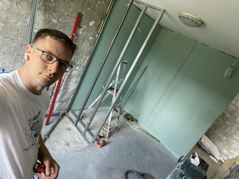 Mężczyzna w okularach wykonujący prace remontowe w pomieszczeniu. W tle zamontowane profile do wykończenia wnętrz oraz narzędzia budowlane, co świadczy o działalności firmy remontowej.