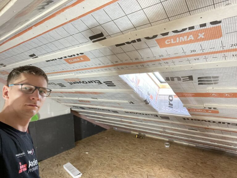 Mężczyzna na zdjęciu wykonuje remont poddasza, przygotowując przestrzeń do wykończenia wnętrz. Na ścianach widoczna izolacja, co wskazuje na profesjonalne usługi firmy remontowej.
