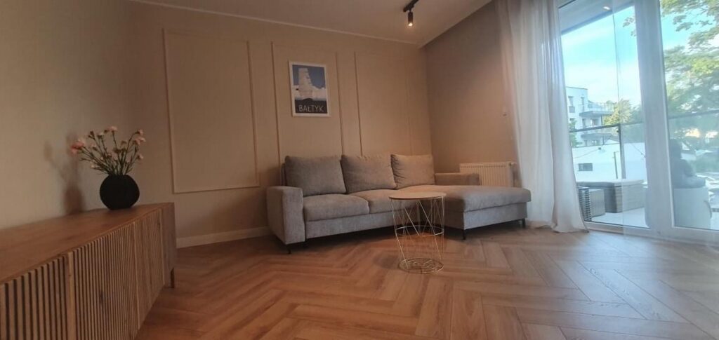 Przodkowo - Nowoczesny salon po remoncie, z eleganckimi wykończeniami wnętrz. Jasne kolory i drewniane podłogi podkreślają styl firm remontowych. W tle duże okno wpuszczające naturalne światło.